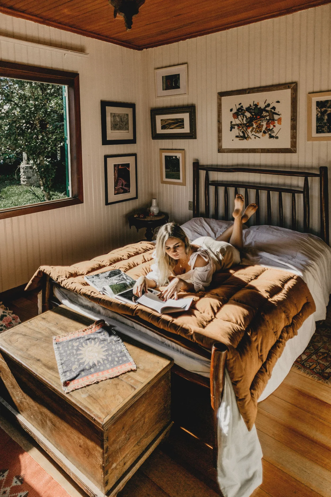 A woman reading a book on a bed in a warmly lit bedroom.