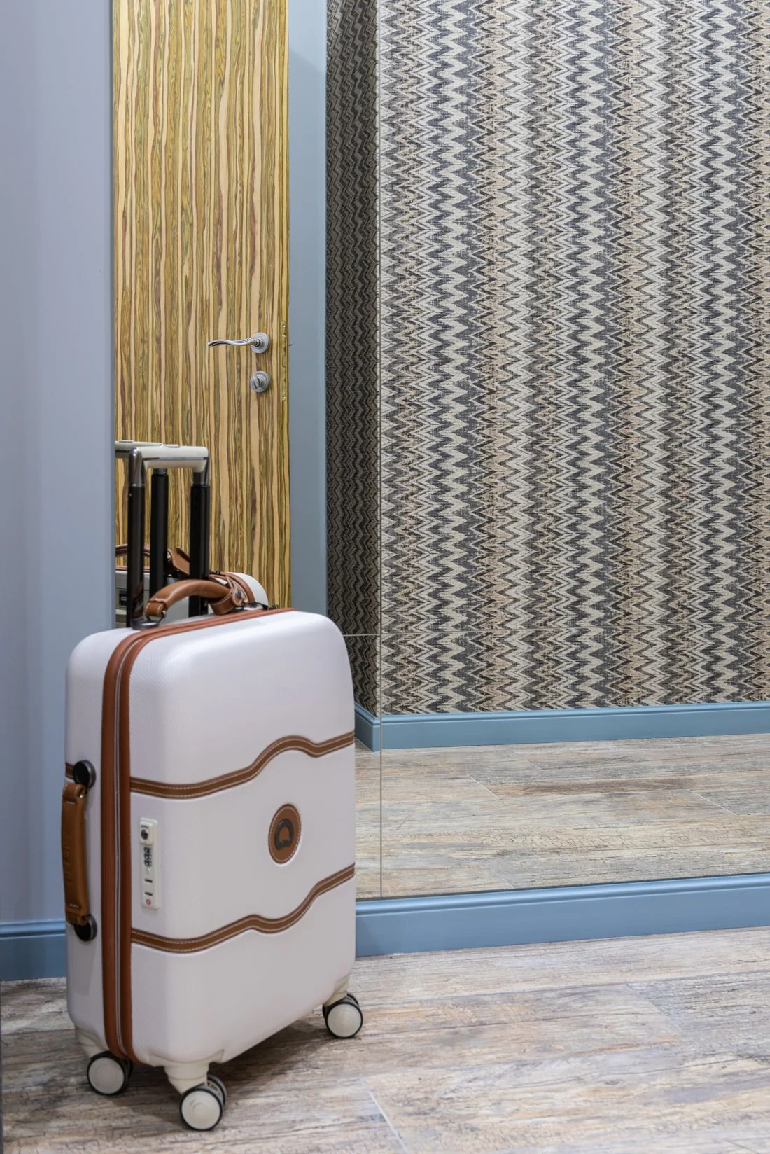 A white suitcase in a hotel corridor beside a mirror.