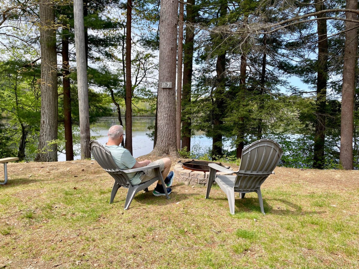 Man sitting in gray Adirondack chair in front of firing overlooking water with pine trees in front