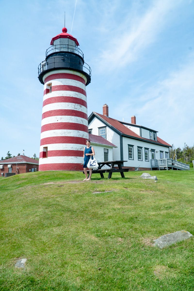 Woman on blue dress holding tote bag with a blue anchor standing in front of red and white striped West Quoddy Head Lighthouse