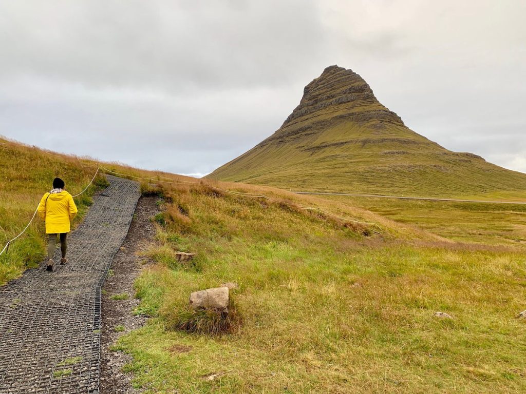 Woman in yellow raincoat walking up path by Kirkjufell